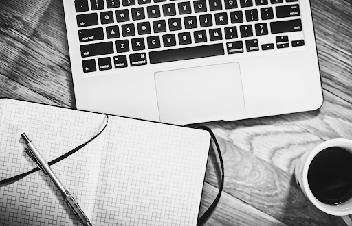 A black-and-white photo of an open notebook, pen, and cup of coffee in front of a laptop.